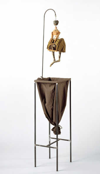 a skeleton figure with a cage like pelvic region, suspended by a hook, hanging over a cloth sack