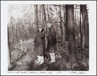 An image of two women in overcoats, wearing hats, gazing sorrowfully at the ground.