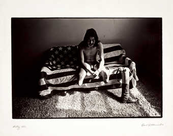 An image of a nude double leg amputee sitting on an American flag draped couch.